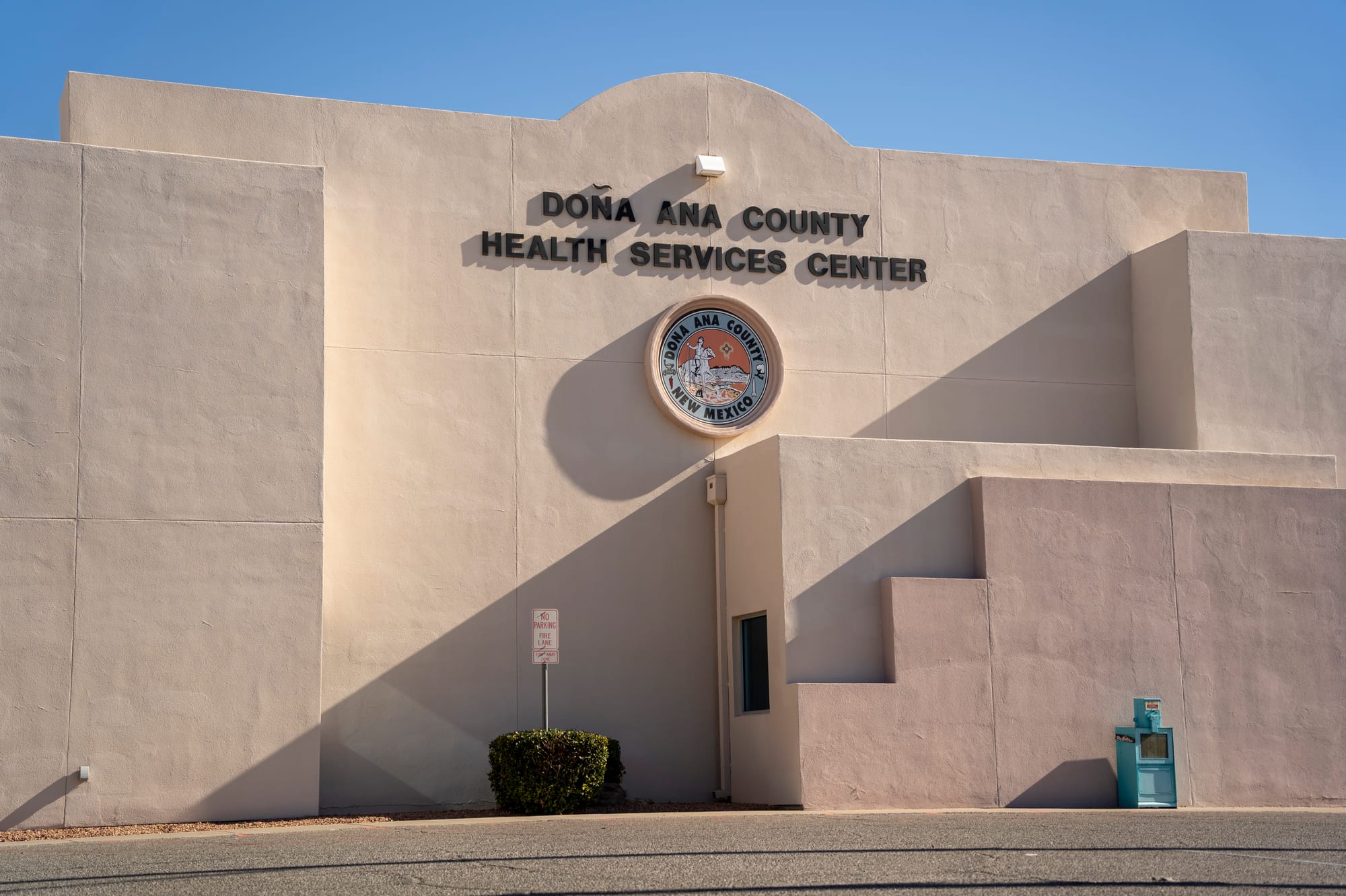 Exterior of the Doña Ana County Health Services Center building in Las Cruces, with the county seal mounted on the stucco facade beneath a clear blue sky.