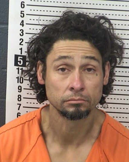 Booking photo of Aaron Trujillo wearing an orange jail uniform in front of a height chart
