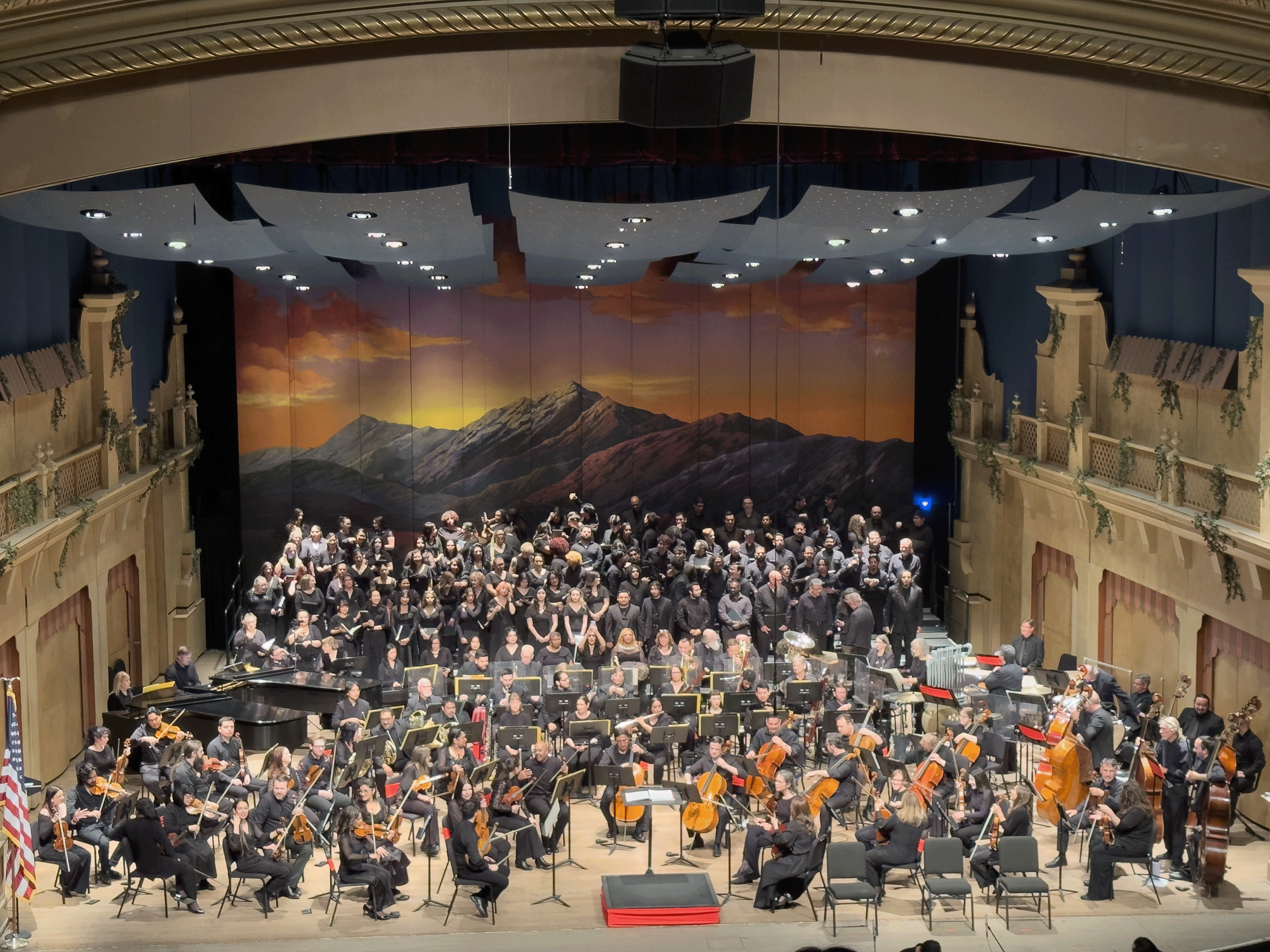 Full orchestra and large choir perform on stage at the Plaza Theatre with a mountain backdrop.