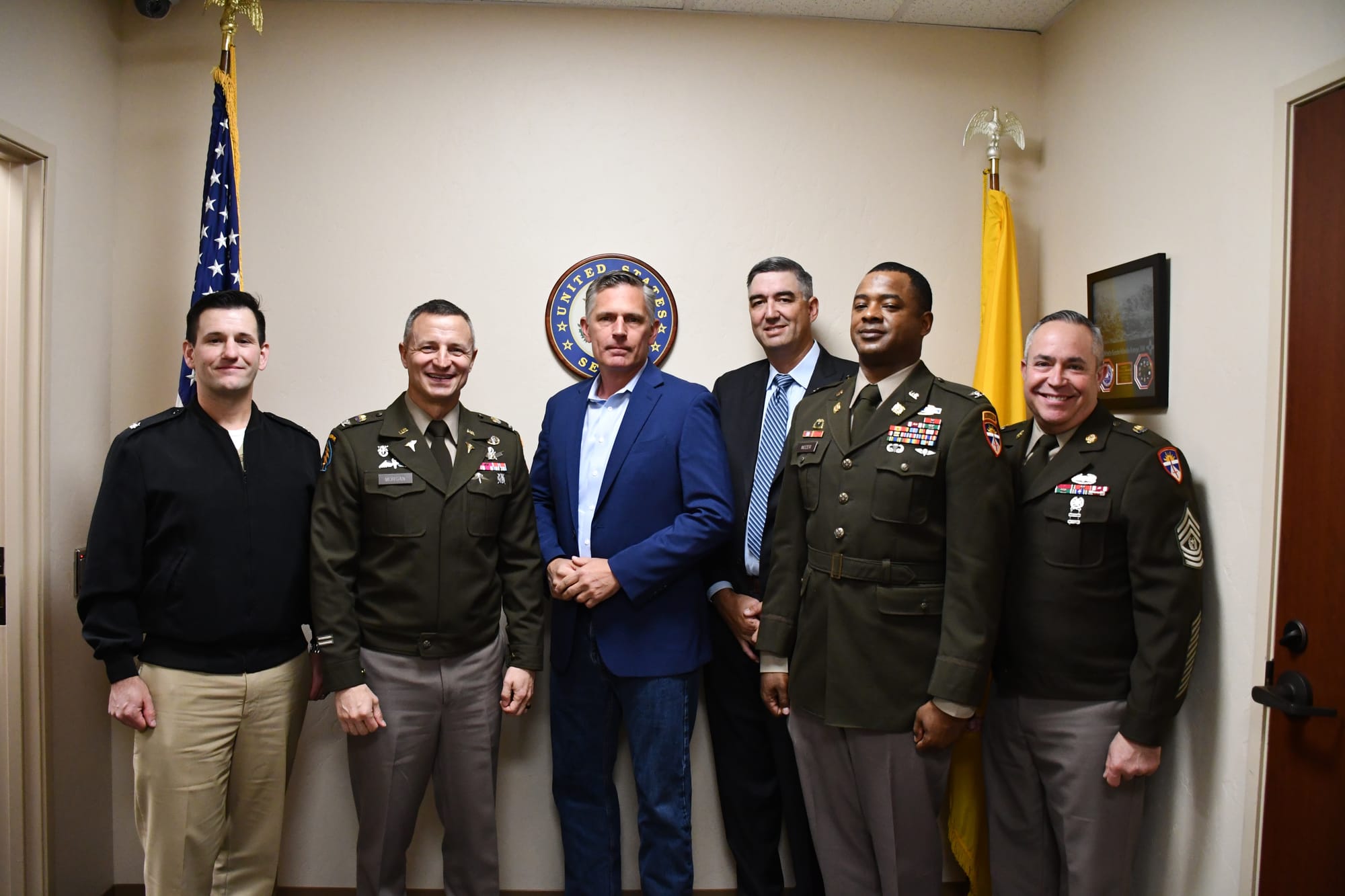 U.S. Sen. Martin Heinrich stands with White Sands Missile Range leadership and military personnel in a group photo inside an office, with U.S. and New Mexico flags in the background.
