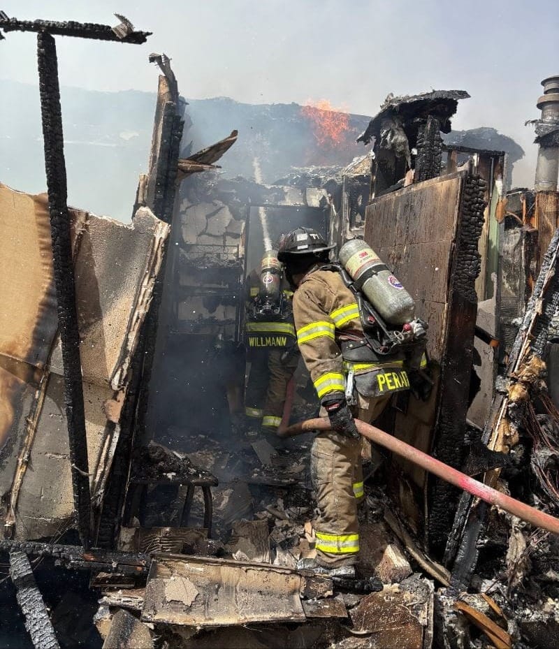 Firefighters in protective gear spray water inside a burned home on Hondo Road, with charred walls, debris and flames still visible.