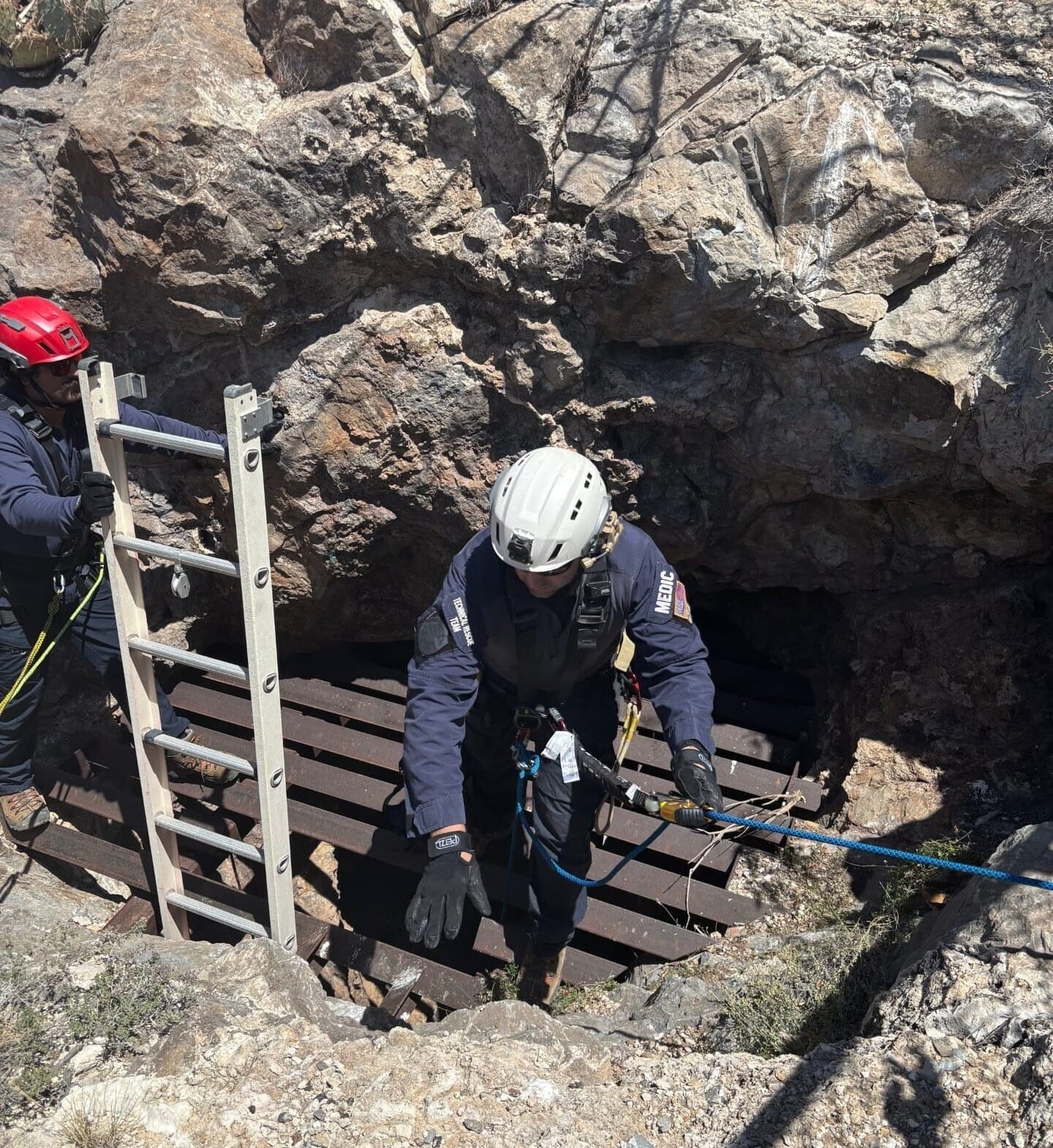 Firefighters wearing helmets and harnesses stand at the edge of a rocky mine shaft, using ropes and a ladder during a rescue operation in the Organ Mountains.