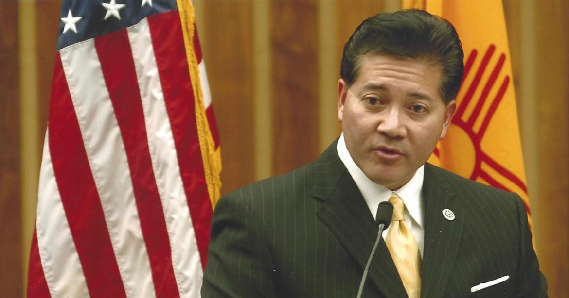 Ken Miyagishima speaks at a podium with U.S. and New Mexico flags visible behind him.