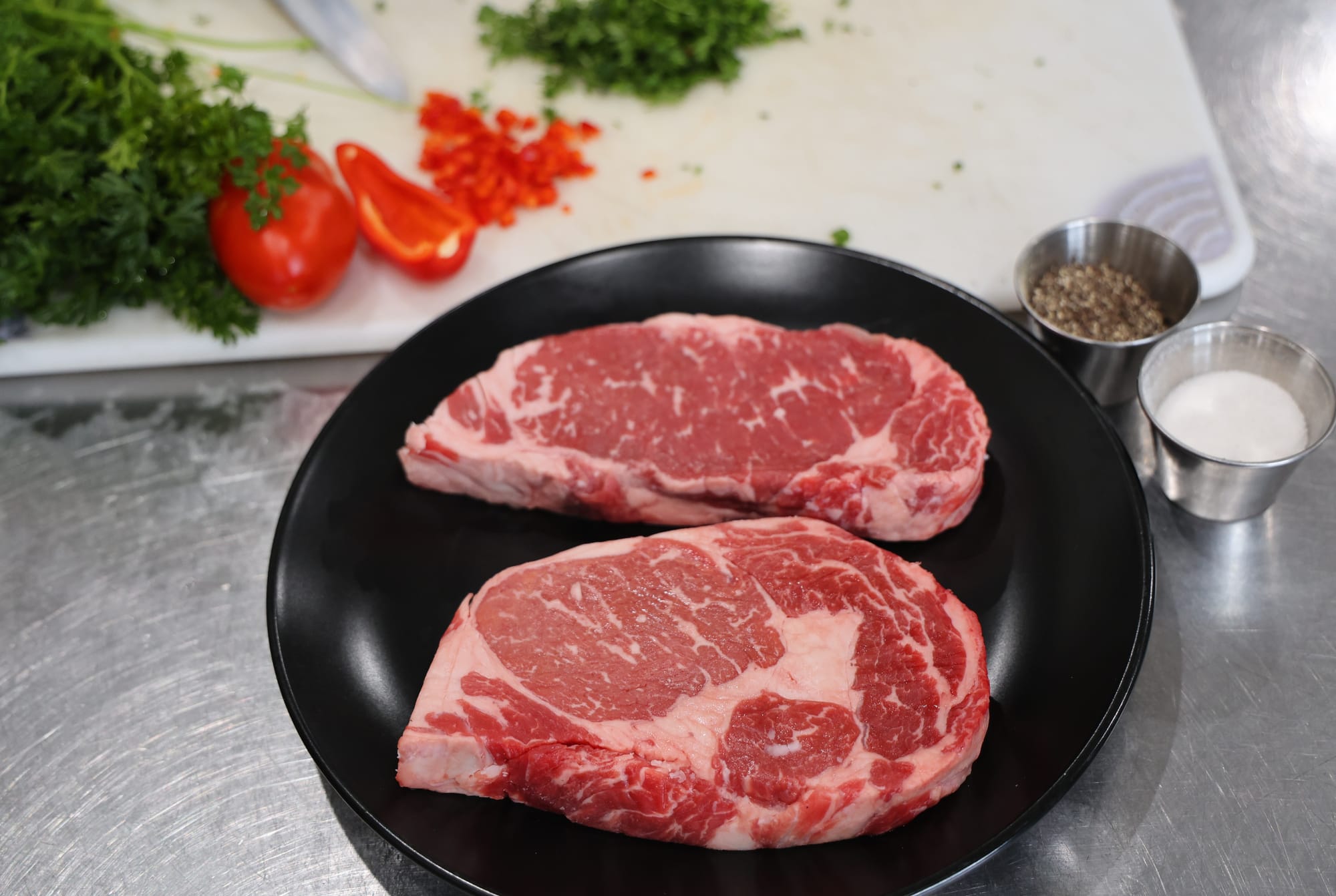 Two raw beef steaks on a plate with chopped vegetables and seasonings on a cutting board, illustrating meat processing and preparation.