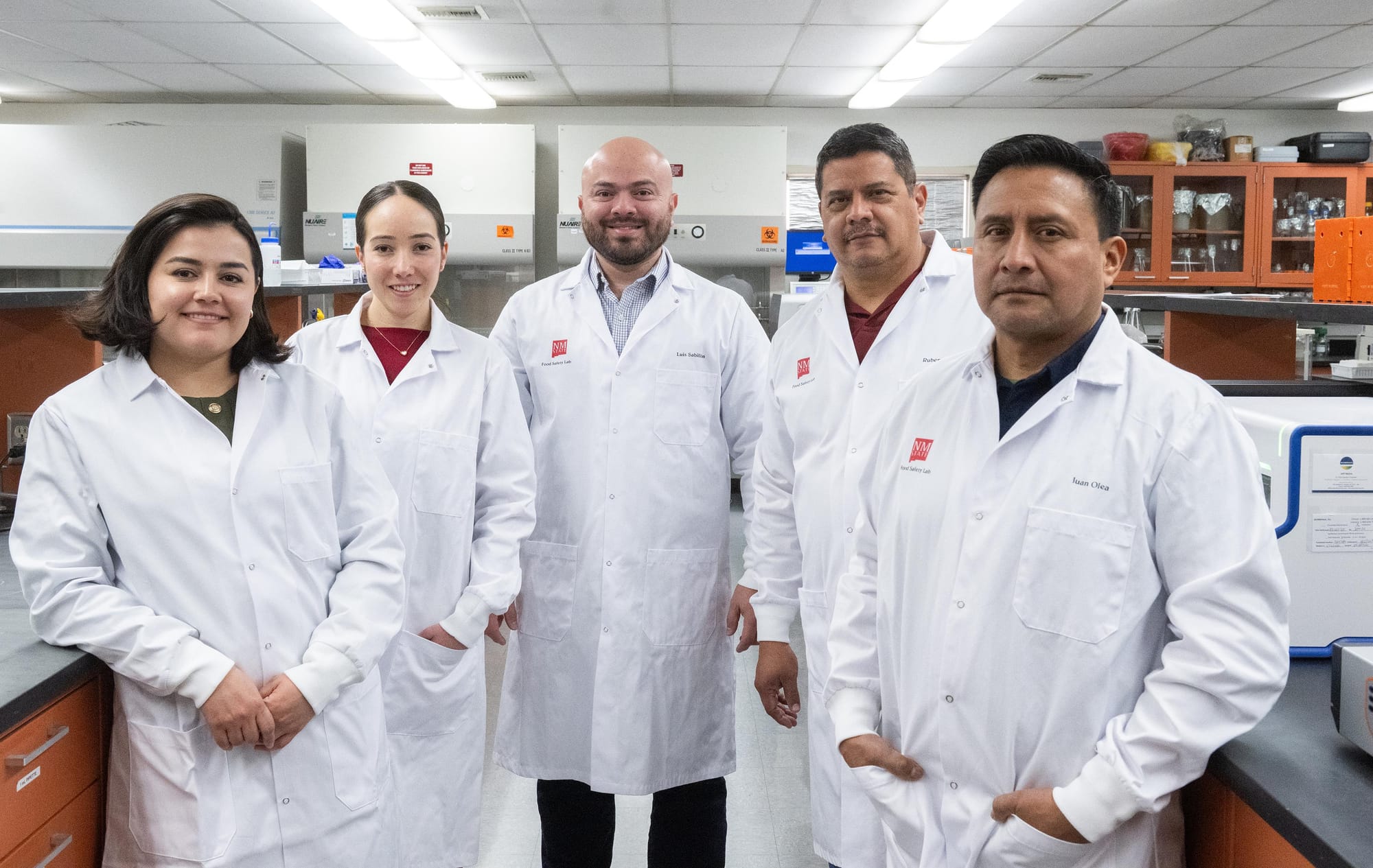 Members of New Mexico State University’s Food Safety Laboratory team stand together in a lab, wearing white lab coats.