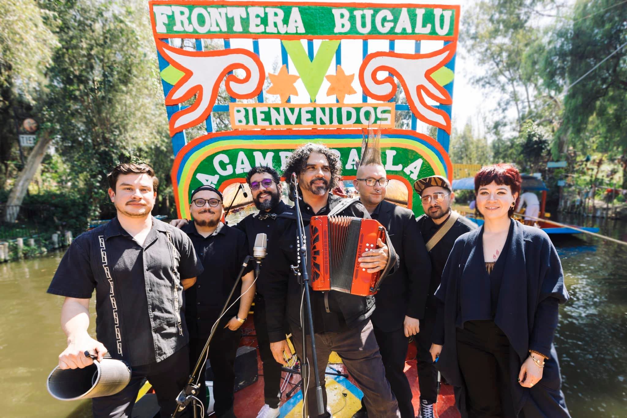 Members of Frontera Bugalú pose together outdoors with instruments in front of a colorful sign.