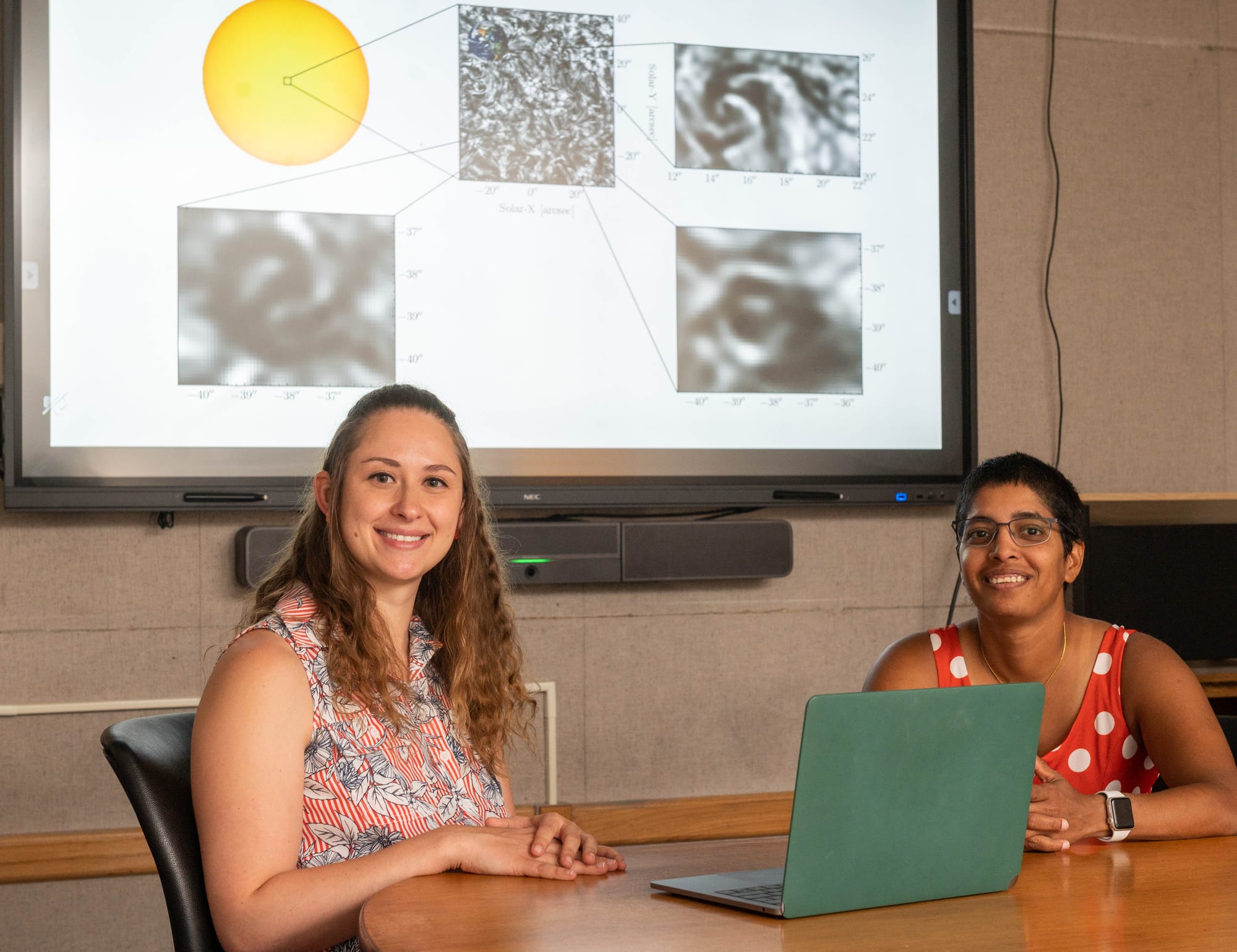 Oana Vesa and Juie Shetye sit at a table with a laptop, with solar research images displayed on a screen behind them.