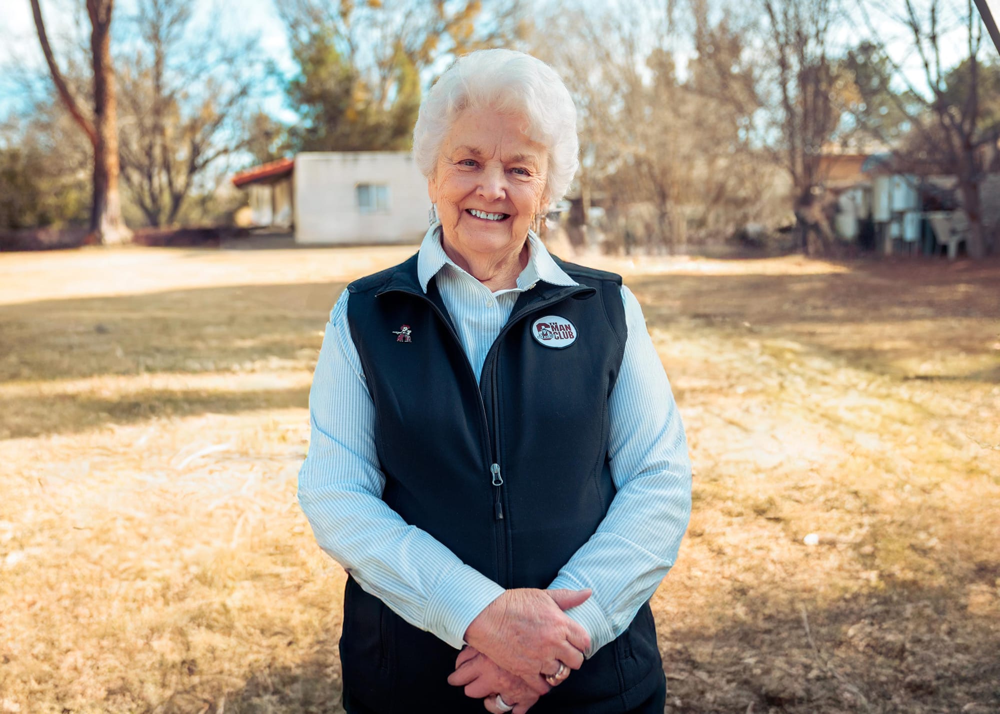 Wanda Bowman stands outdoors smiling in a yard, wearing a vest and collared shirt.