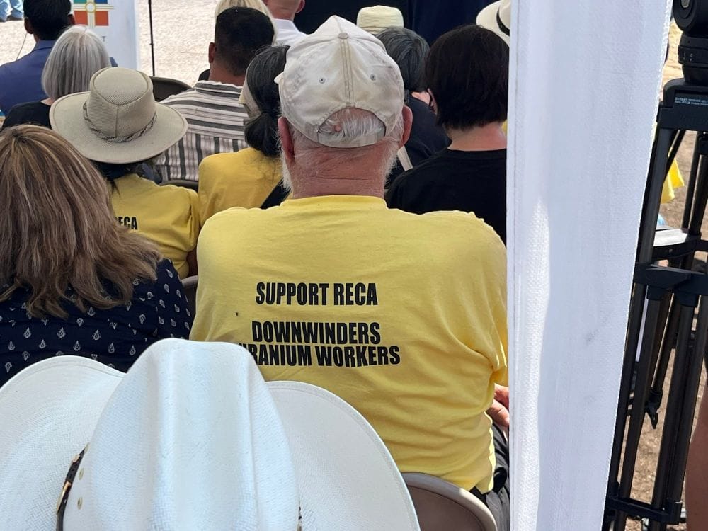 Audience members sit at a RECA event, including a man wearing a yellow shirt that reads “Support RECA Downwinders Uranium Workers.”