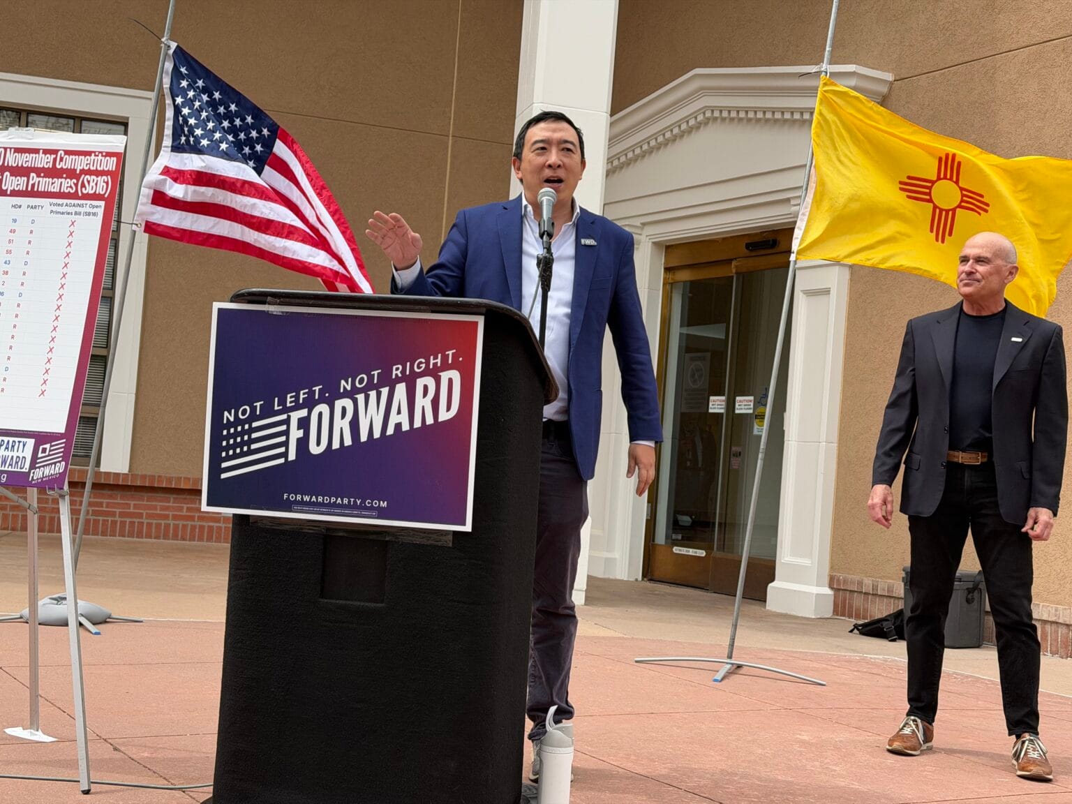 Andrew Yang speaks at a podium during a Forward Party event in New Mexico, with U.S. and state flags displayed behind him.