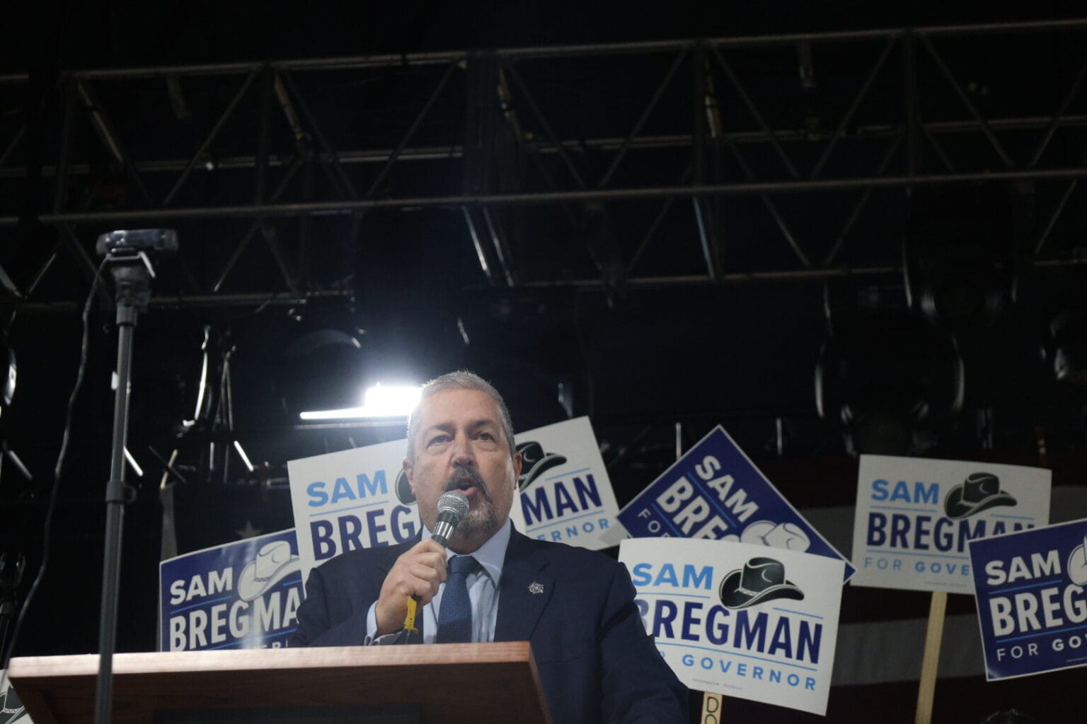 Sam Bregman speaks at a campaign event while supporters hold “Sam Bregman for Governor” signs behind him.