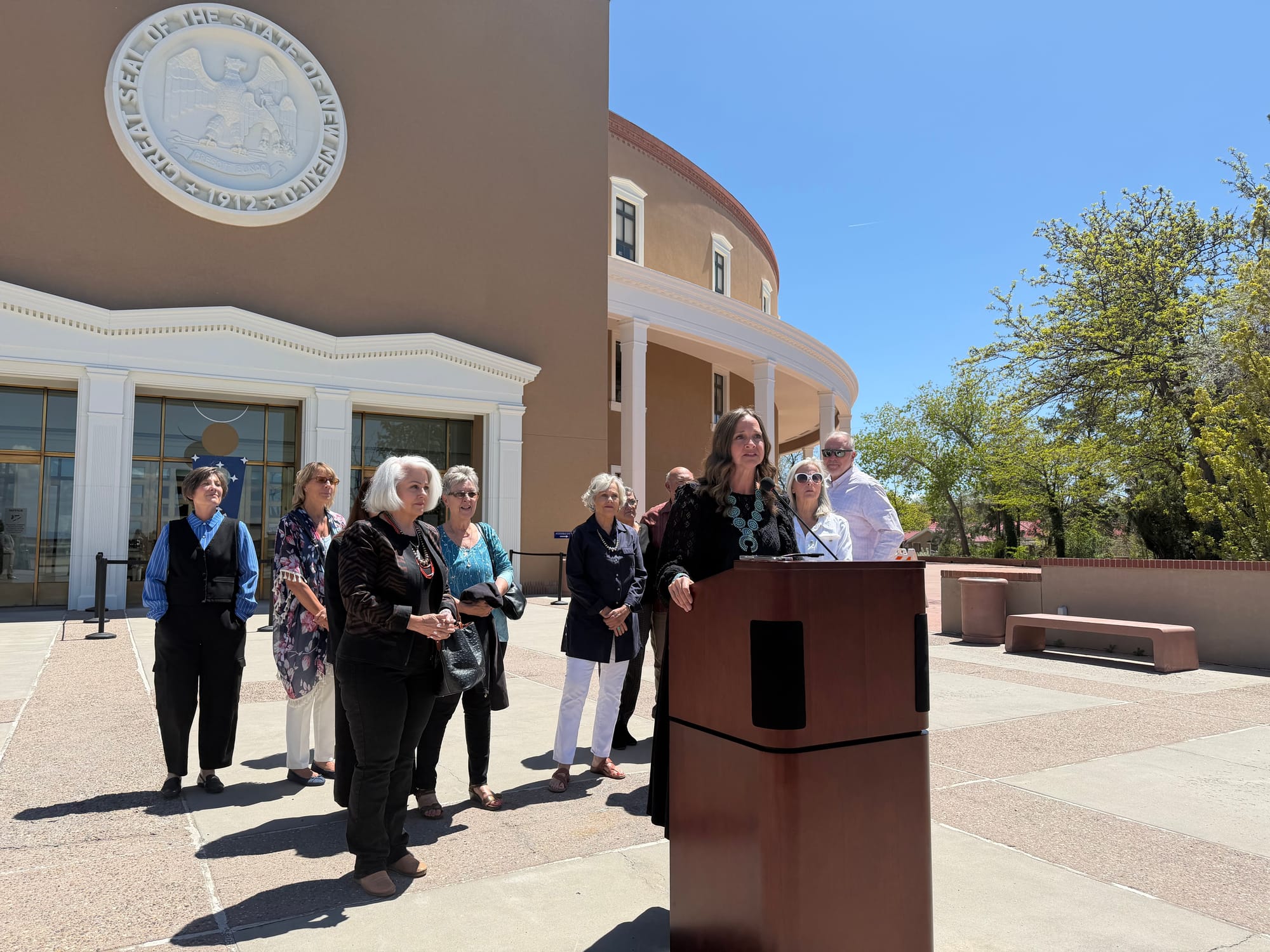 Rep. Rebecca Dow speaks at a podium outside the New Mexico State Capitol, with a group of supporters standing behind her.
