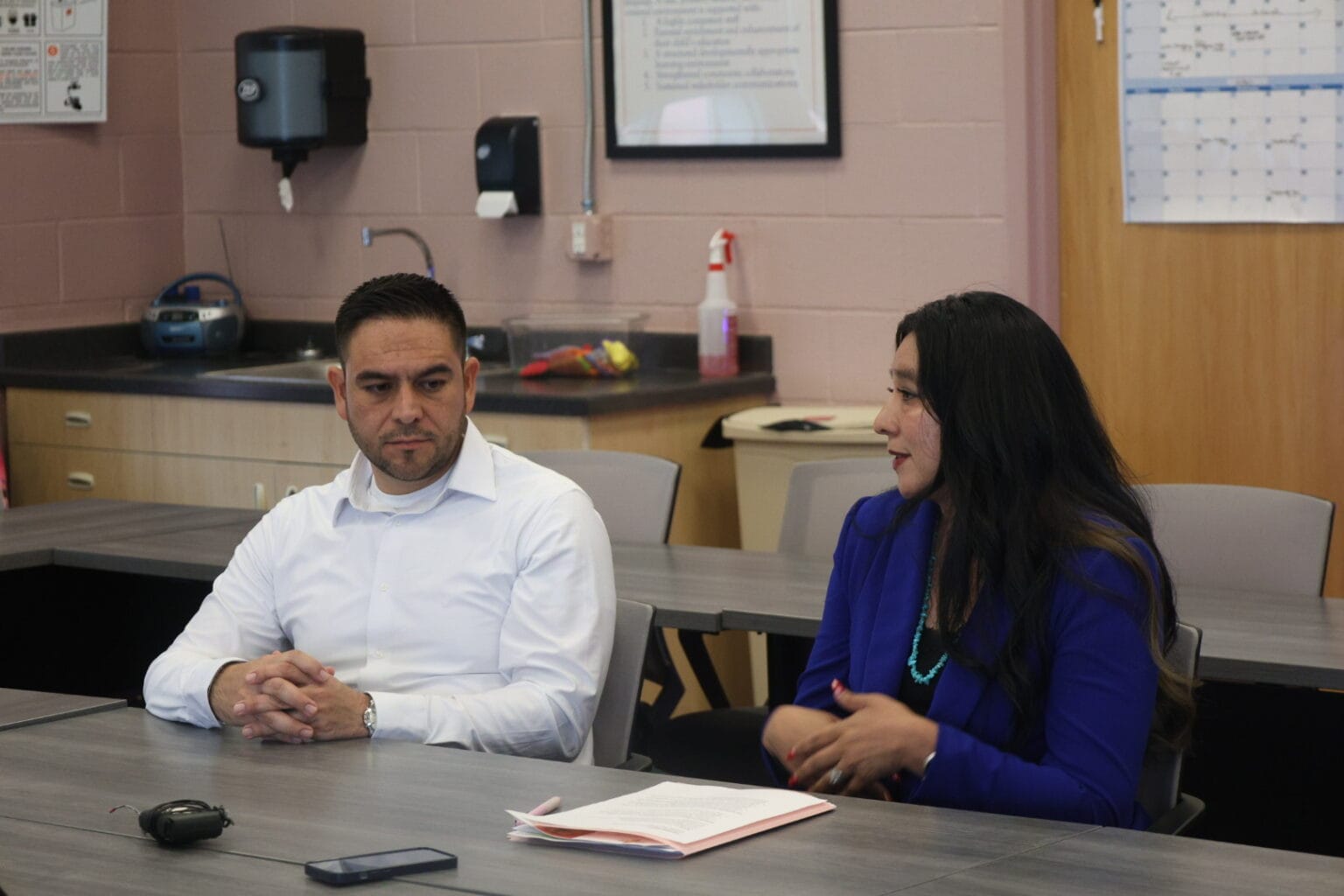 Two people sit at a table during a meeting, with one speaking while the other listens in a classroom or conference room setting.