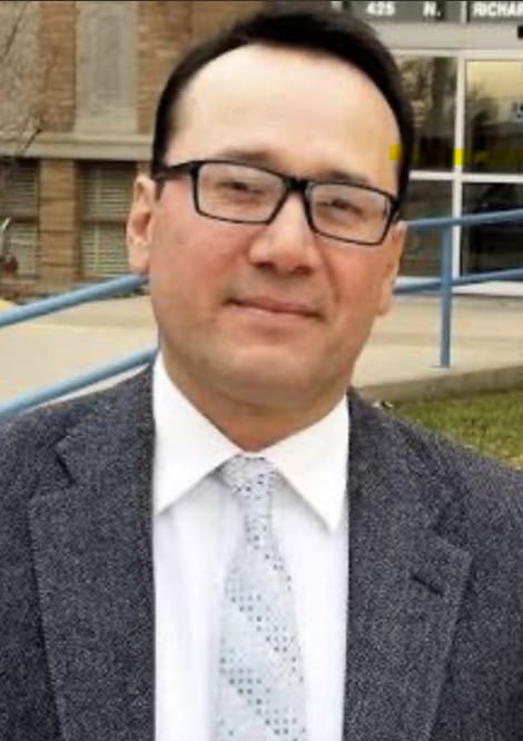 Juan Fuentes stands outdoors wearing glasses, a suit jacket and tie in front of a building entrance.