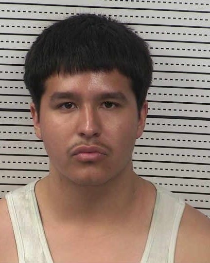 Booking photo of a man with short dark hair and a mustache wearing a light-colored tank top, facing the camera against a height chart background.