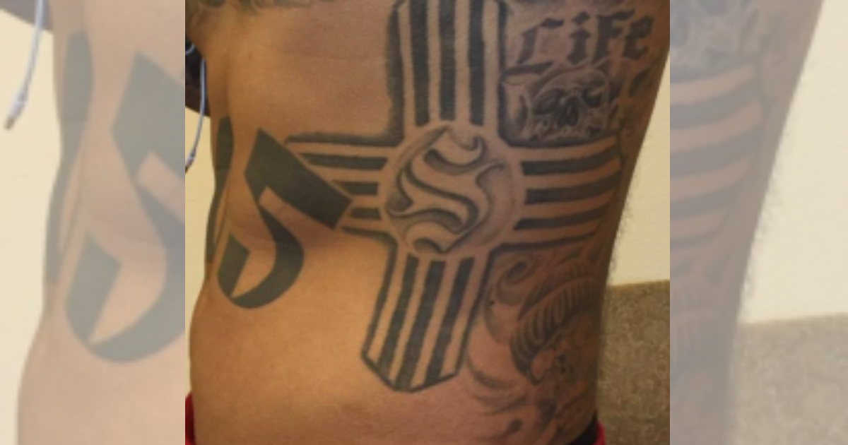 Close-up of a torso tattoo featuring a stylized Zia symbol associated with the Syndicato de Nuevo Mexico gang.