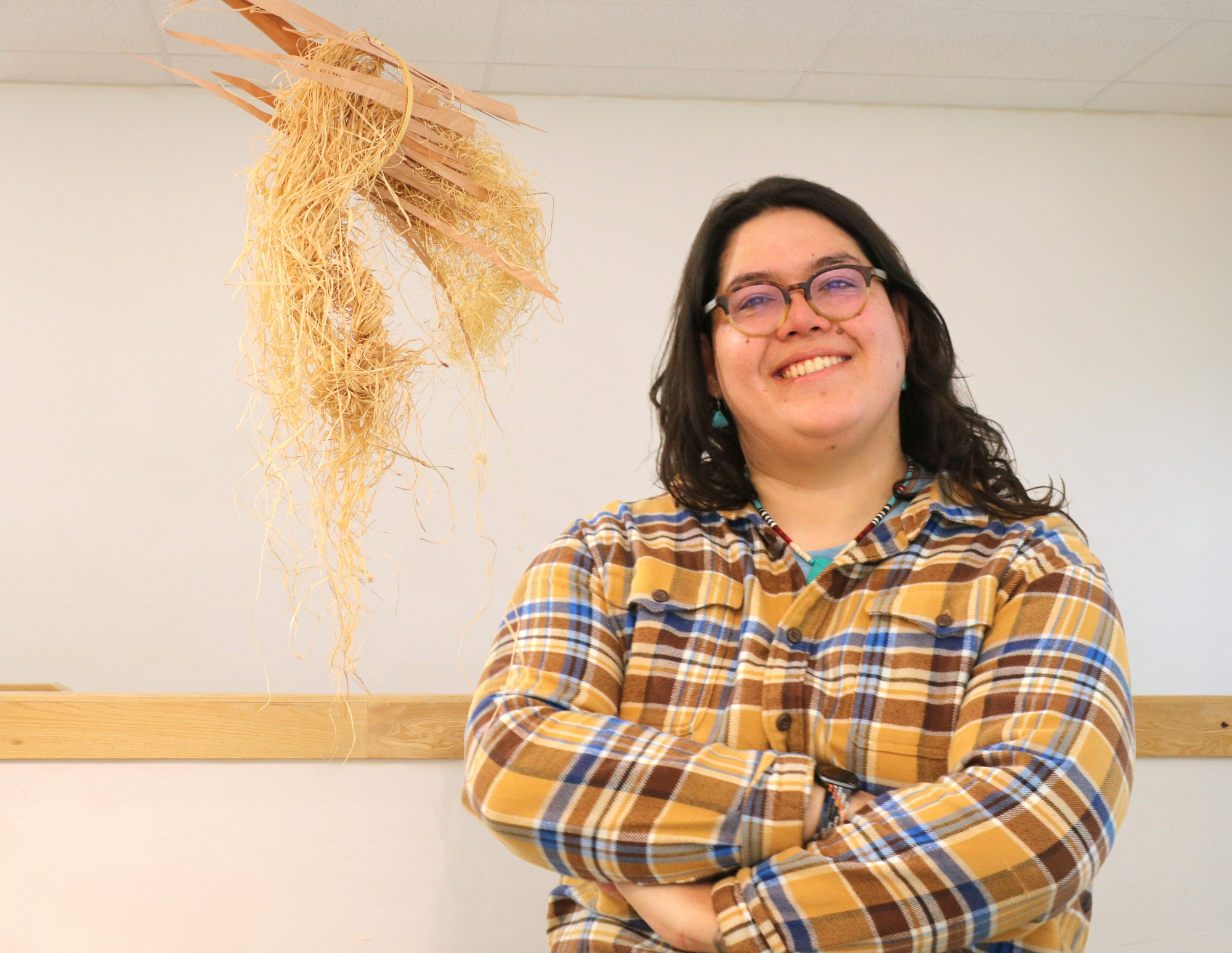 Renee Maderazo smiles with arms crossed beside a hanging fiber sculpture made of raffia in an indoor studio space.