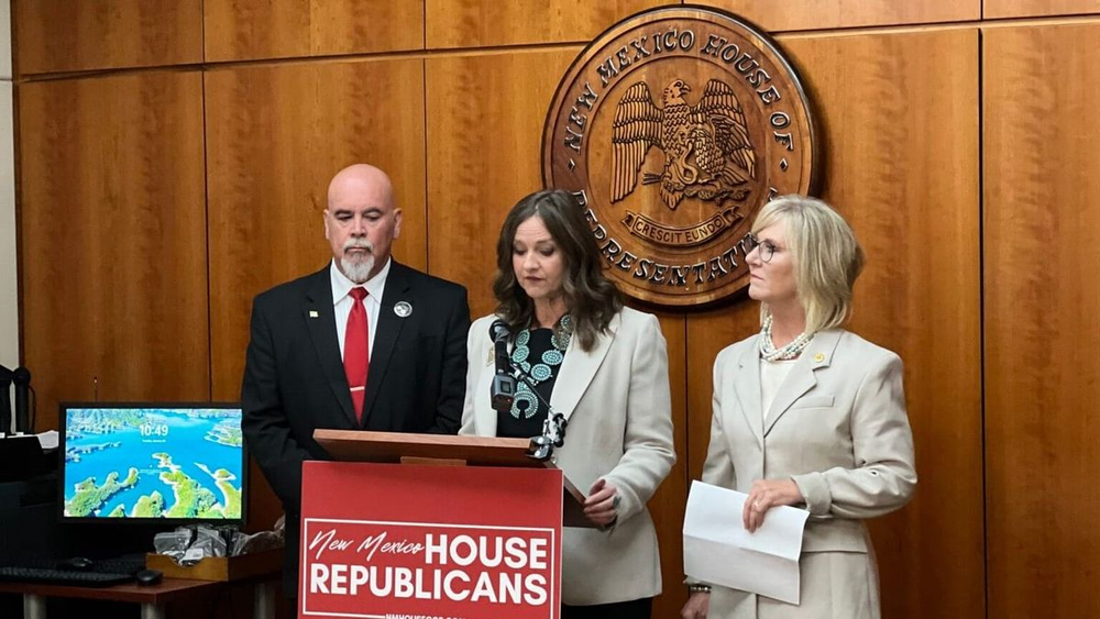 New Mexico state Rep. Rebecca Dow speaks at a podium during a news conference at the Roundhouse, flanked by two colleagues with the New Mexico House of Representatives seal visible behind them.