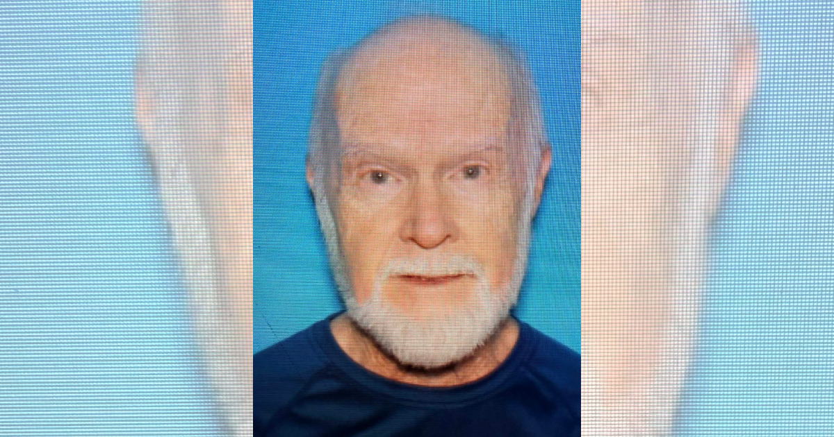 Photo of 85-year-old George Douglas Alexander, a missing Las Cruces man with short gray hair and a white beard, wearing a dark shirt against a blue background.