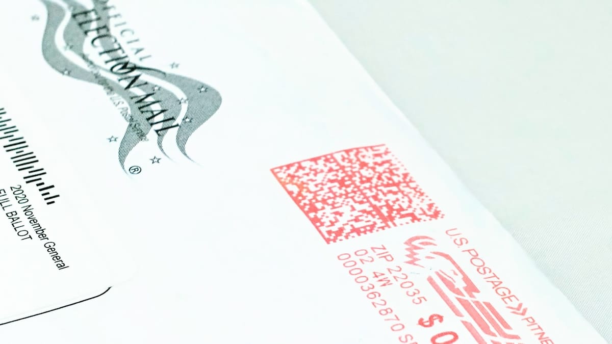 Close-up of a mail ballot envelope with U.S. Postal Service postage markings and a barcode.