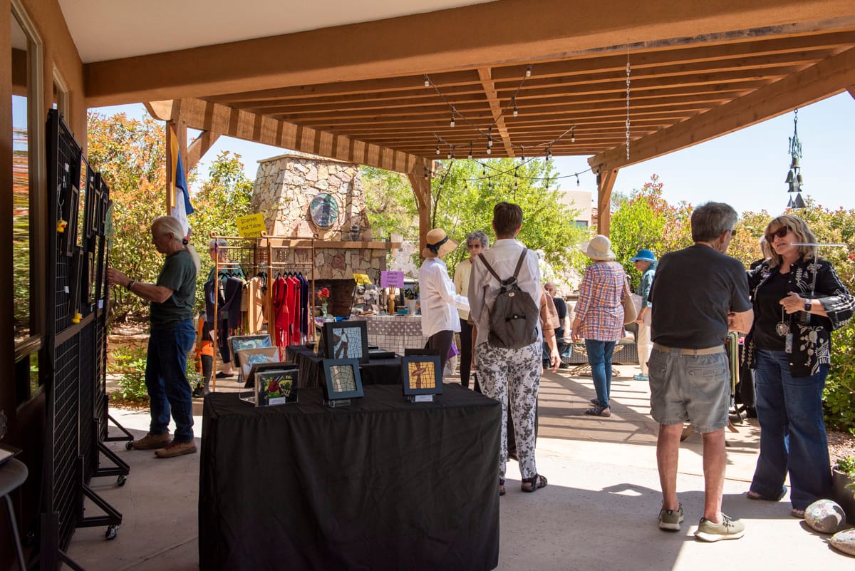Visitors browse artwork and interact with vendors at an outdoor Art in the Garden event under a covered patio in Picacho Hills.