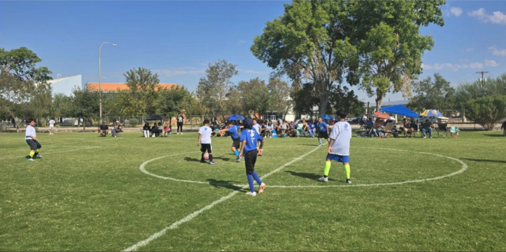 Children play soccer on a grassy field during a youth outdoor league game in Las Cruces while spectators watch from the sidelines.