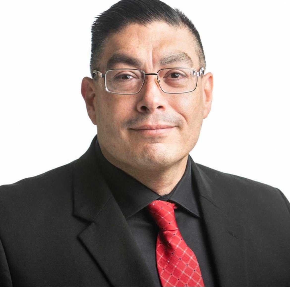 Republican Doña Ana County sheriff candidate Jaime Quezada poses for a portrait wearing glasses, a black suit and red tie.