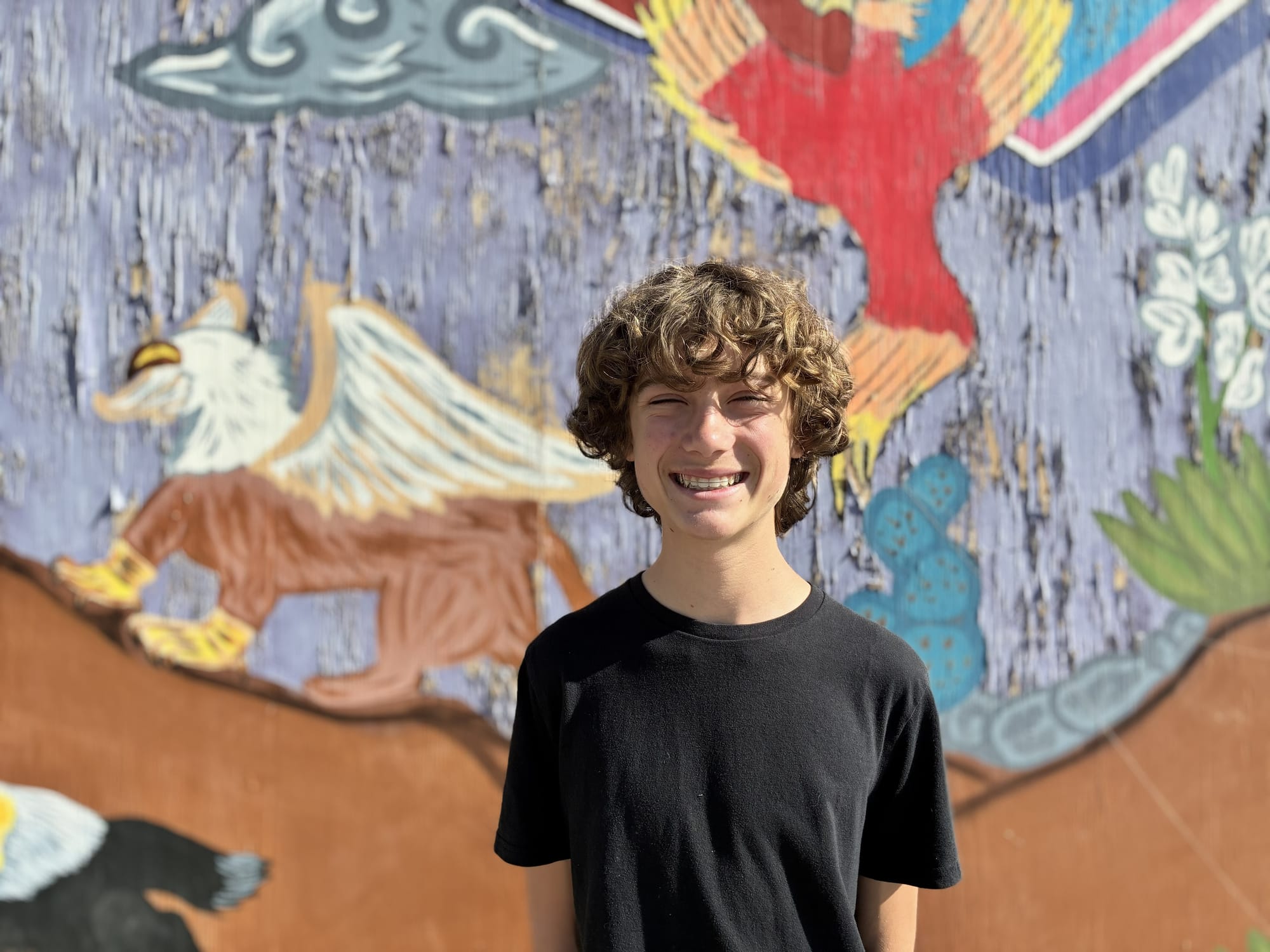 Mesilla Valley Leadership Academy student Adam Cram stands smiling in front of a colorful mural.