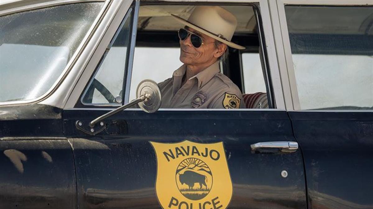 Zahn McClarnon, wearing a Navajo police uniform, sunglasses and cowboy hat, sits inside a Navajo Police vehicle in a scene from AMC’s “Dark Winds.”