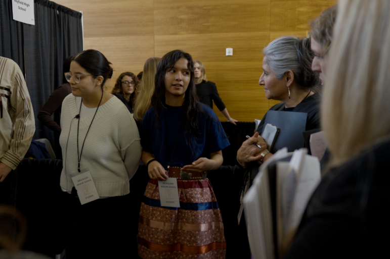 Student speaks with an adult judge during a STEM competition while other participants stand nearby.