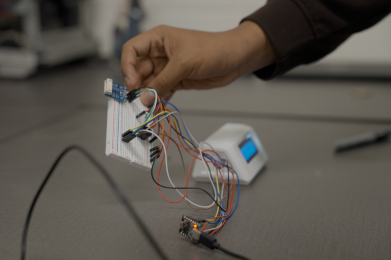 Hand holds a small electronic sensor device with wires connected to a circuit board on a tabletop.
