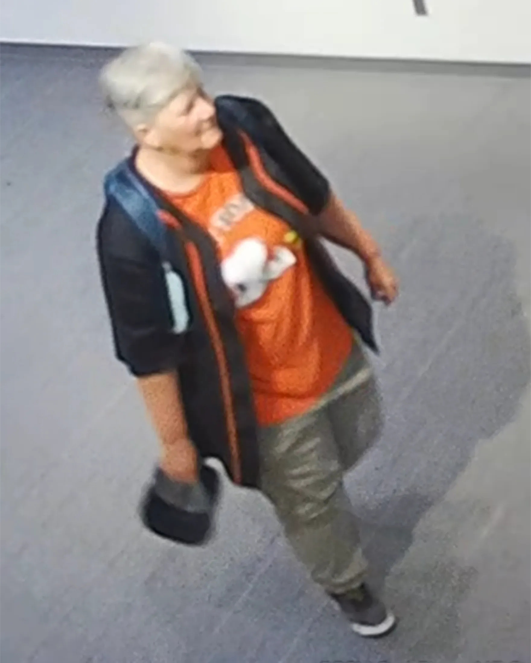 Surveillance image of a woman walking indoors, wearing an orange shirt and dark jacket, released by NMSU Police as part of an arson investigation.