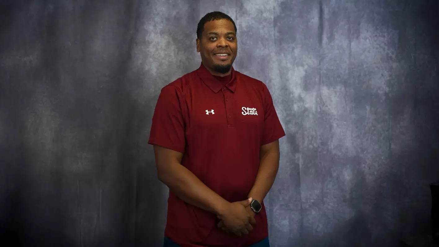 Brandon Barnes stands in a red NM State polo shirt during a posed portrait photo against a gray studio backdrop.
