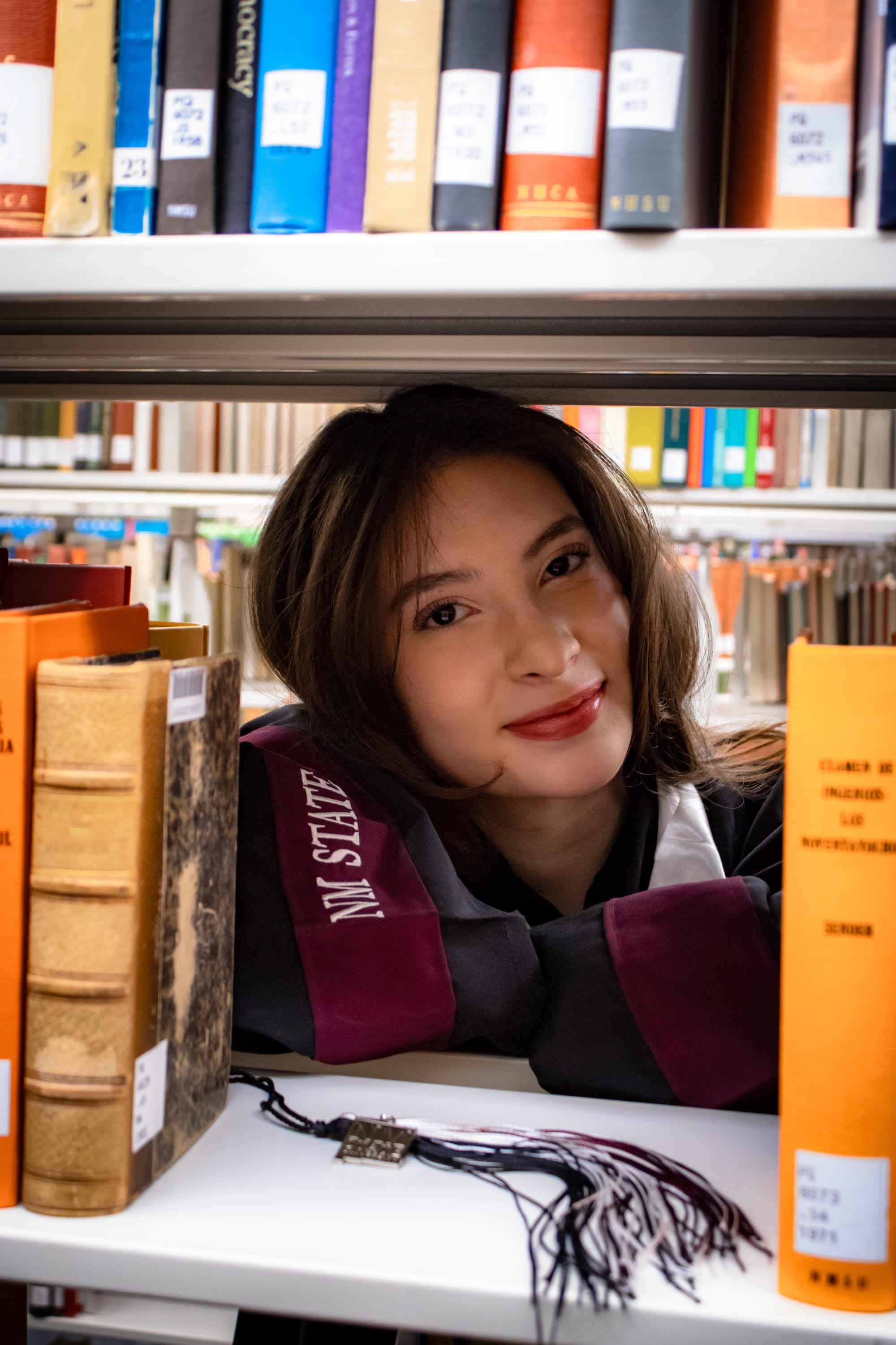 NMSU graduate Genesis Valenzo poses between library shelves while wearing graduation regalia.