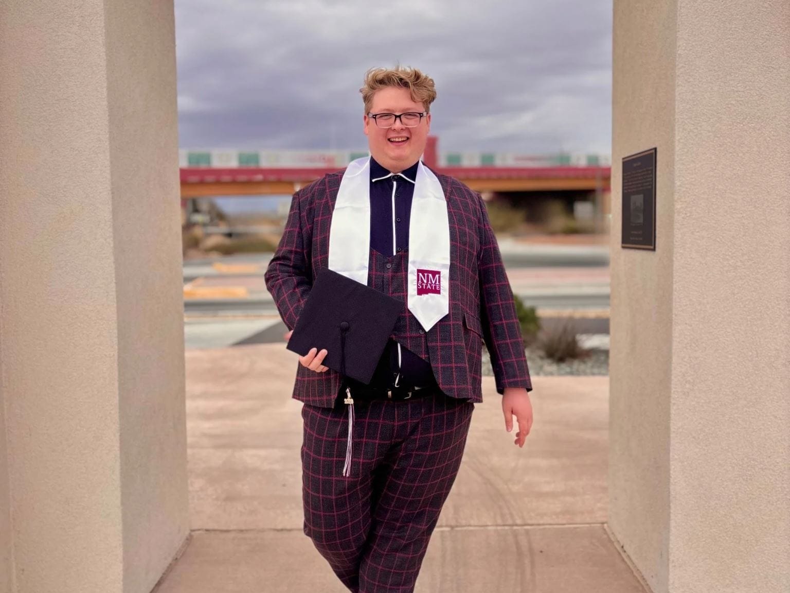 NMSU graduate Atlee Musgrave poses in graduation attire while holding a mortarboard cap outdoors on campus.