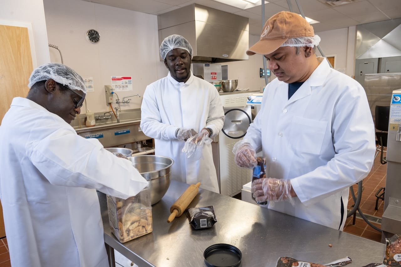 Three New Mexico State University graduate students wearing lab coats, hairnets and gloves prepare ice cream ingredients on a stainless steel worktable in the campus food science dairy lab.