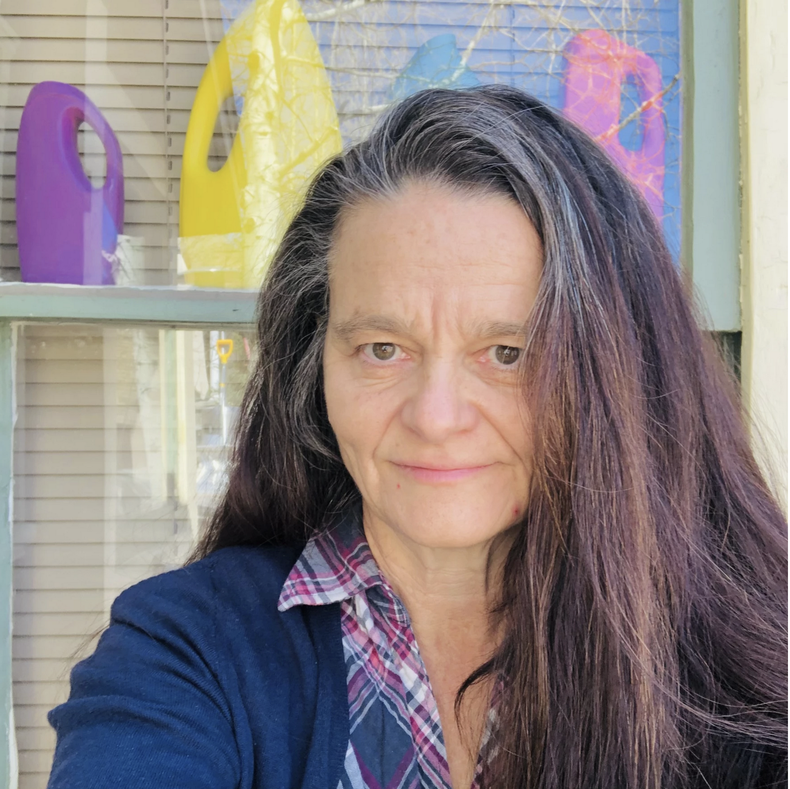 Connie Voisine, a poetry professor at New Mexico State University, stands outdoors in front of a window while wearing a plaid shirt and dark cardigan.