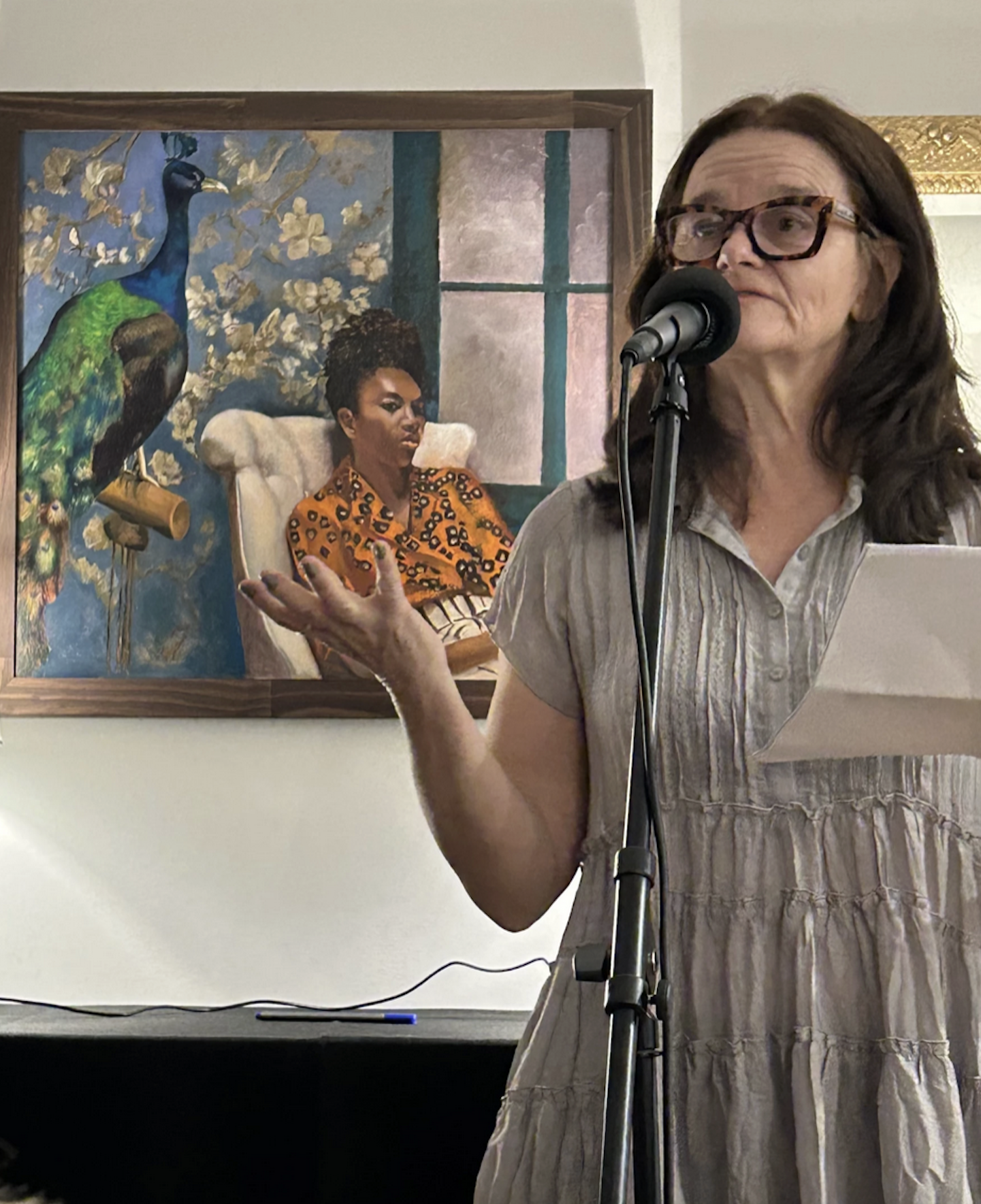 Connie Voisine reads from a sheet of paper while speaking into a microphone during a poetry reading, with a large framed painting visible behind her.
