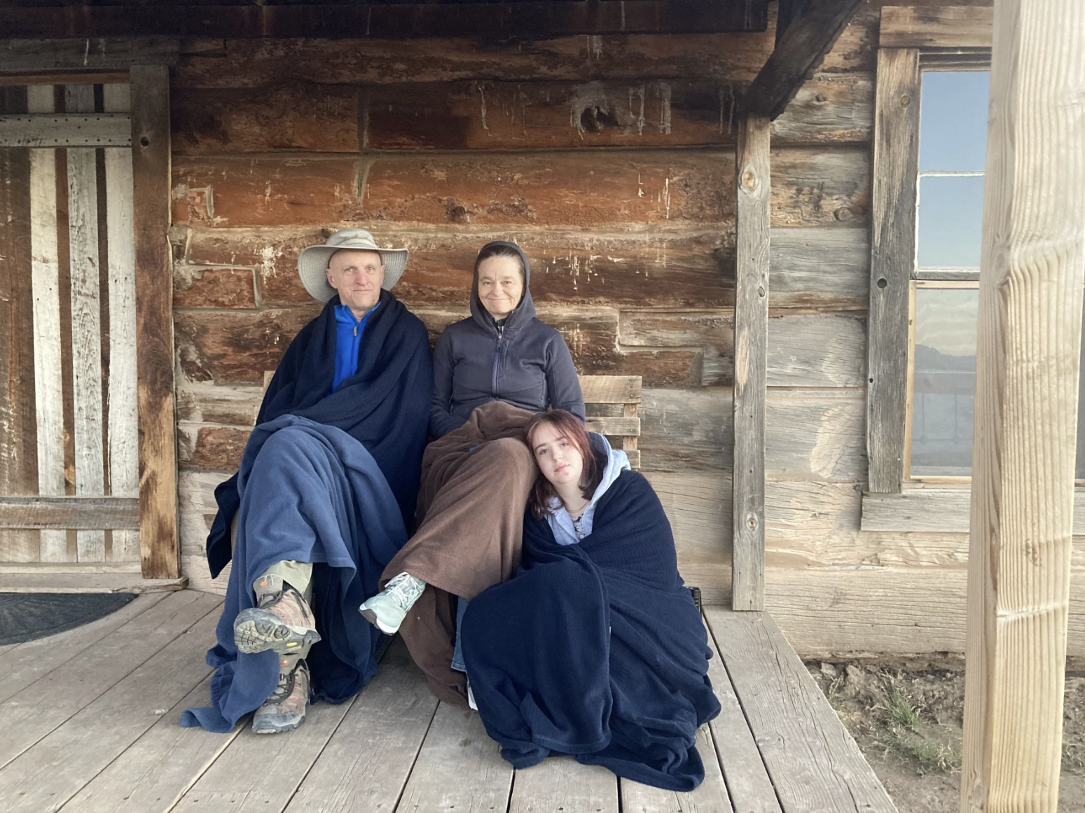 Connie Voisine sits on a wooden porch with her husband, Rus Bradburd, and their daughter, Alma, wrapped in blankets outside a rustic cabin.