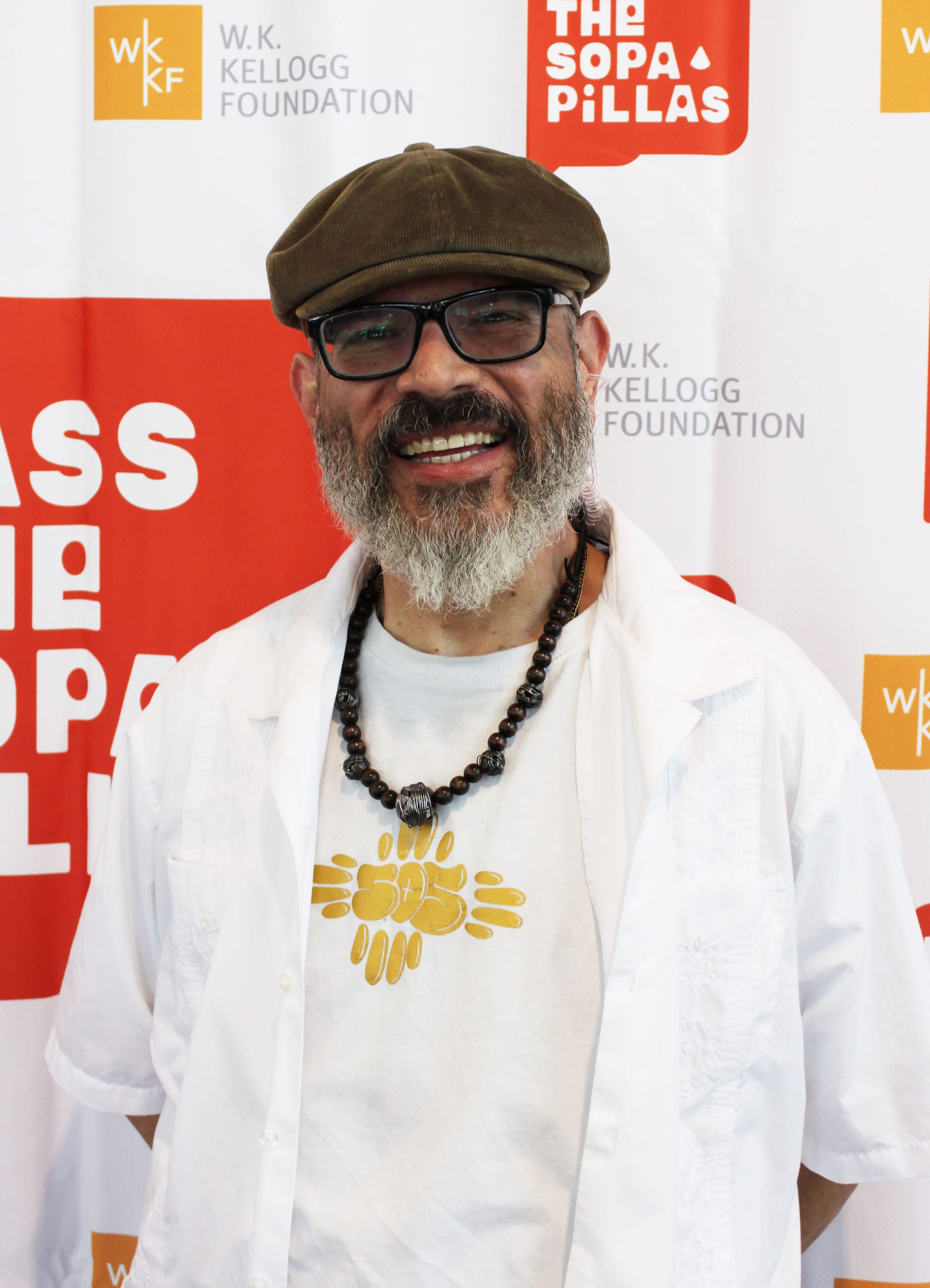 Manuel González, host of the “Pass the Sopapillas” podcast and New Mexico state poet laureate, smiles while standing in front of a W.K. Kellogg Foundation and “Pass the Sopapillas” backdrop.