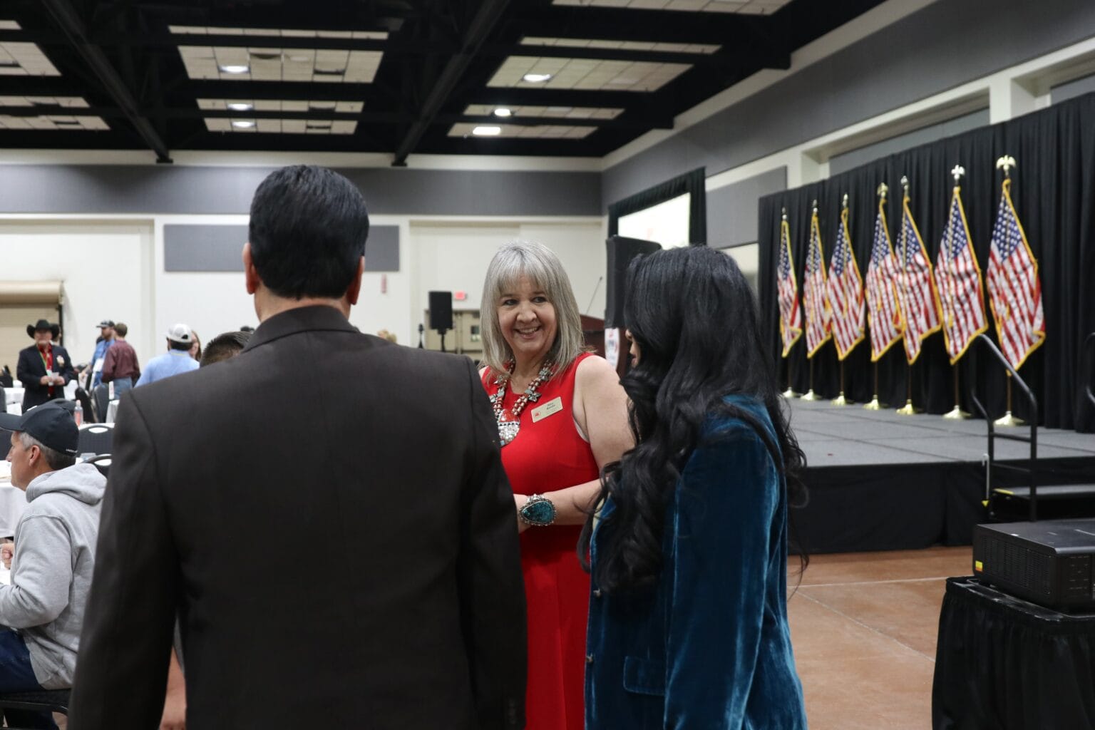 Amy Barela, chair of the New Mexico Republican Party, speaks with attendees at an indoor event with American flags in the background.