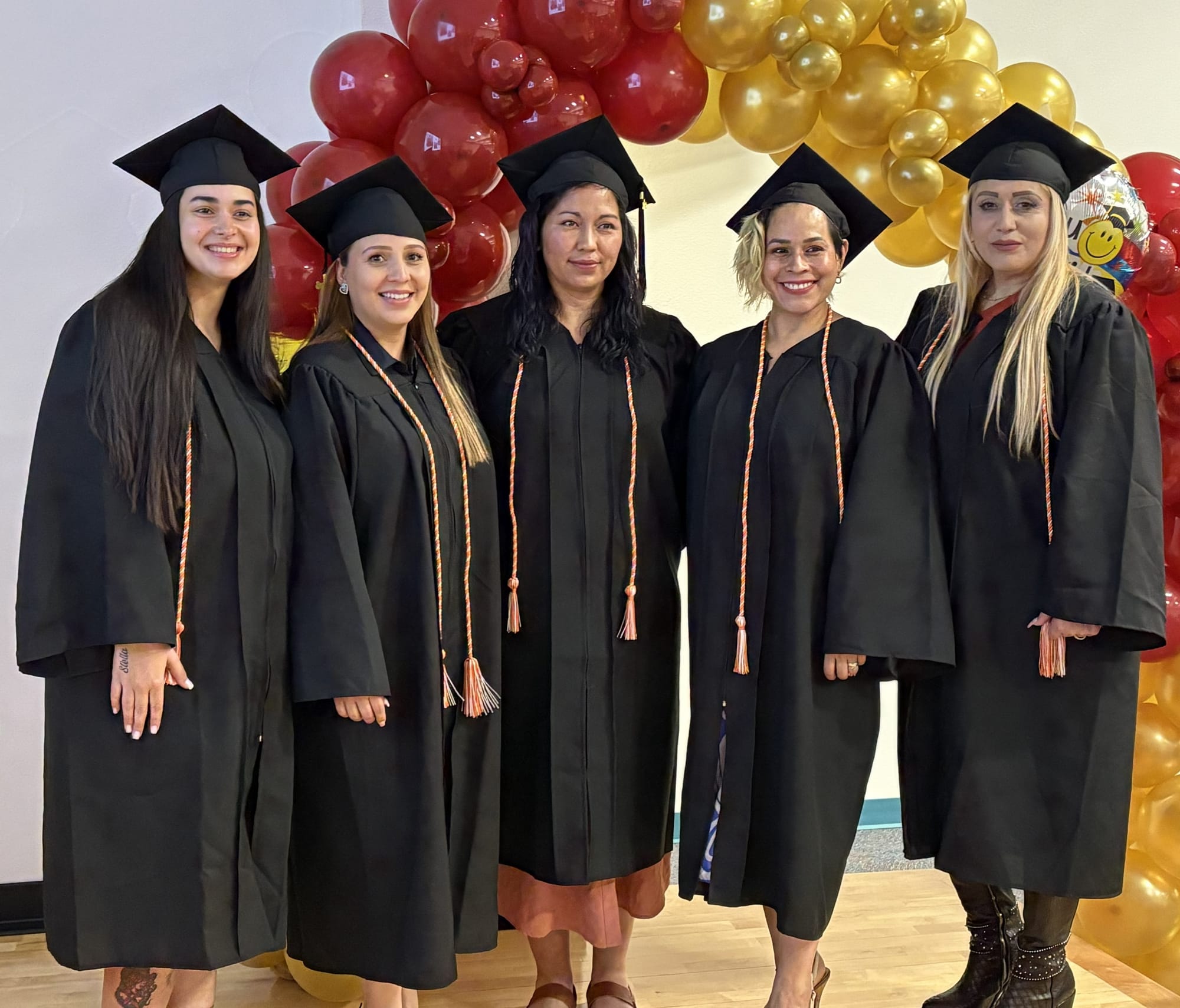 Five AVANCE to College graduates stand in caps and gowns in front of a red and gold balloon arch.
