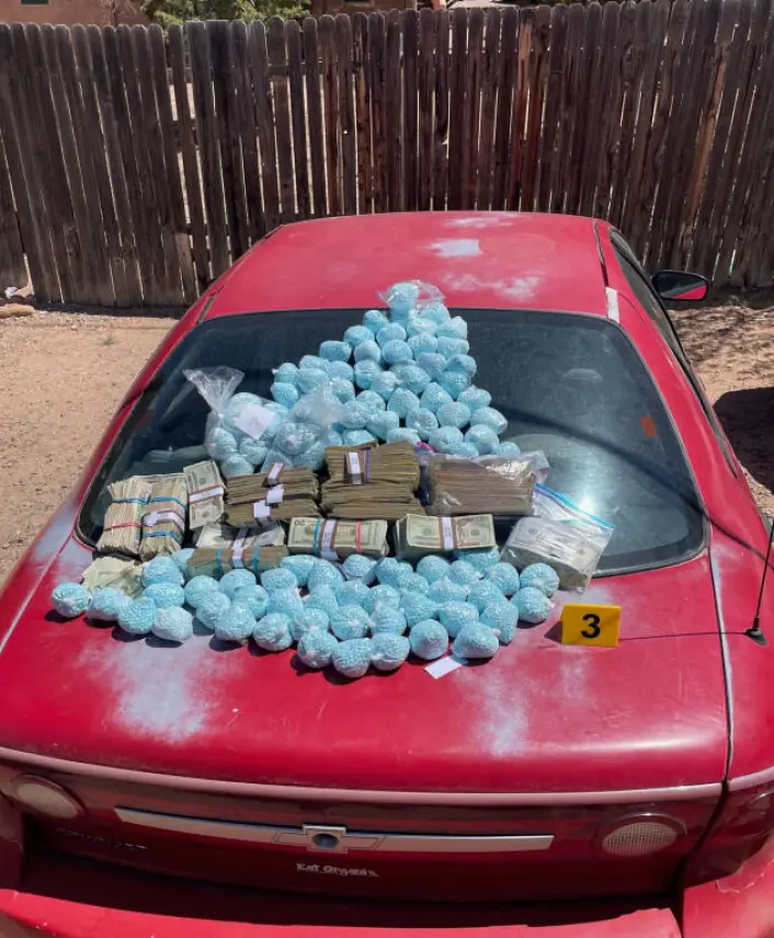 Blue fentanyl pills and stacks of cash are displayed on the rear window and trunk of a red car.