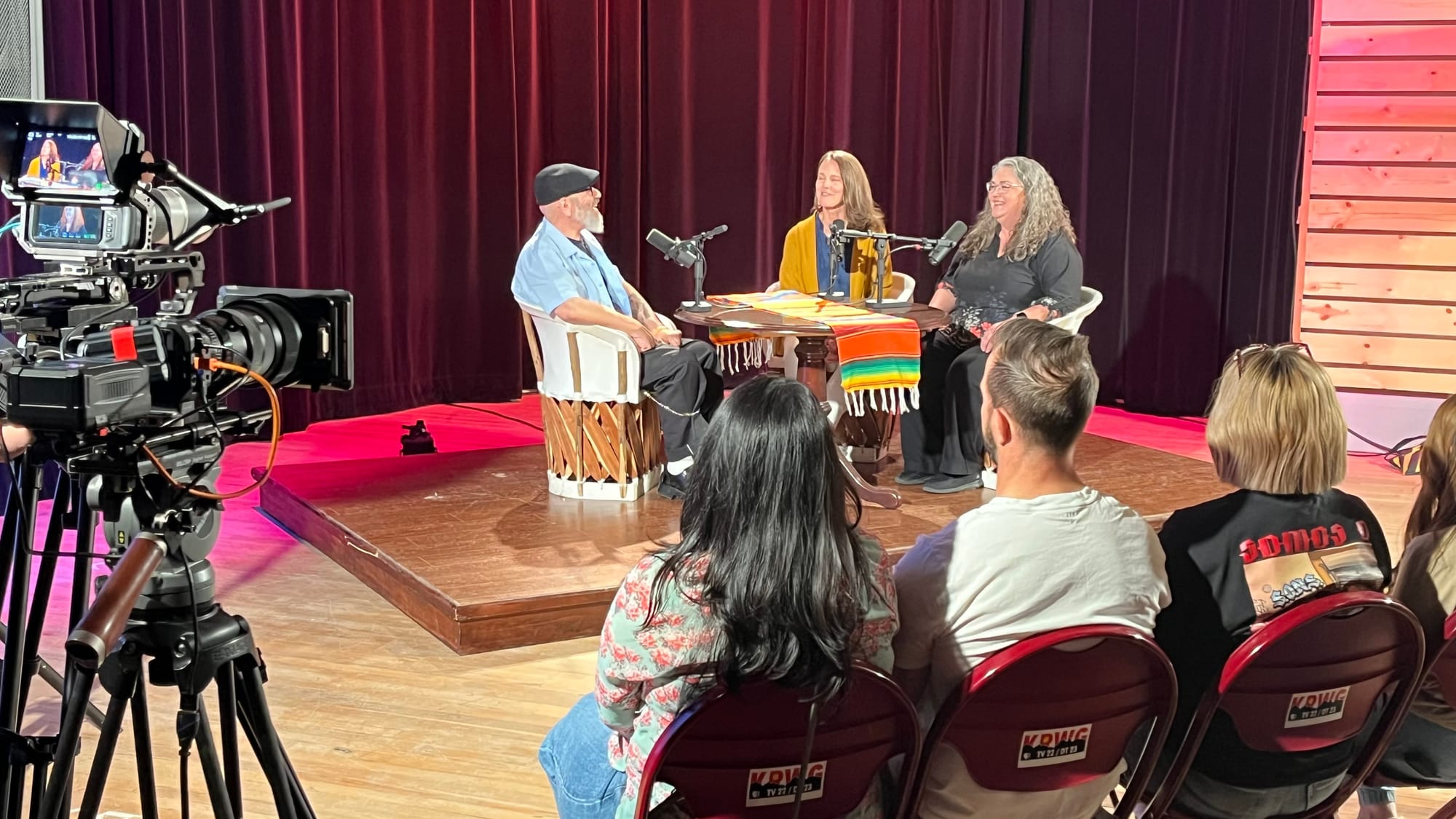 A video camera records Manuel González, seated at left onstage, as he speaks with Terra Winter and Mary Ulrich during a live “Pass the Sopapillas” taping at KRWG Studios before an audience.