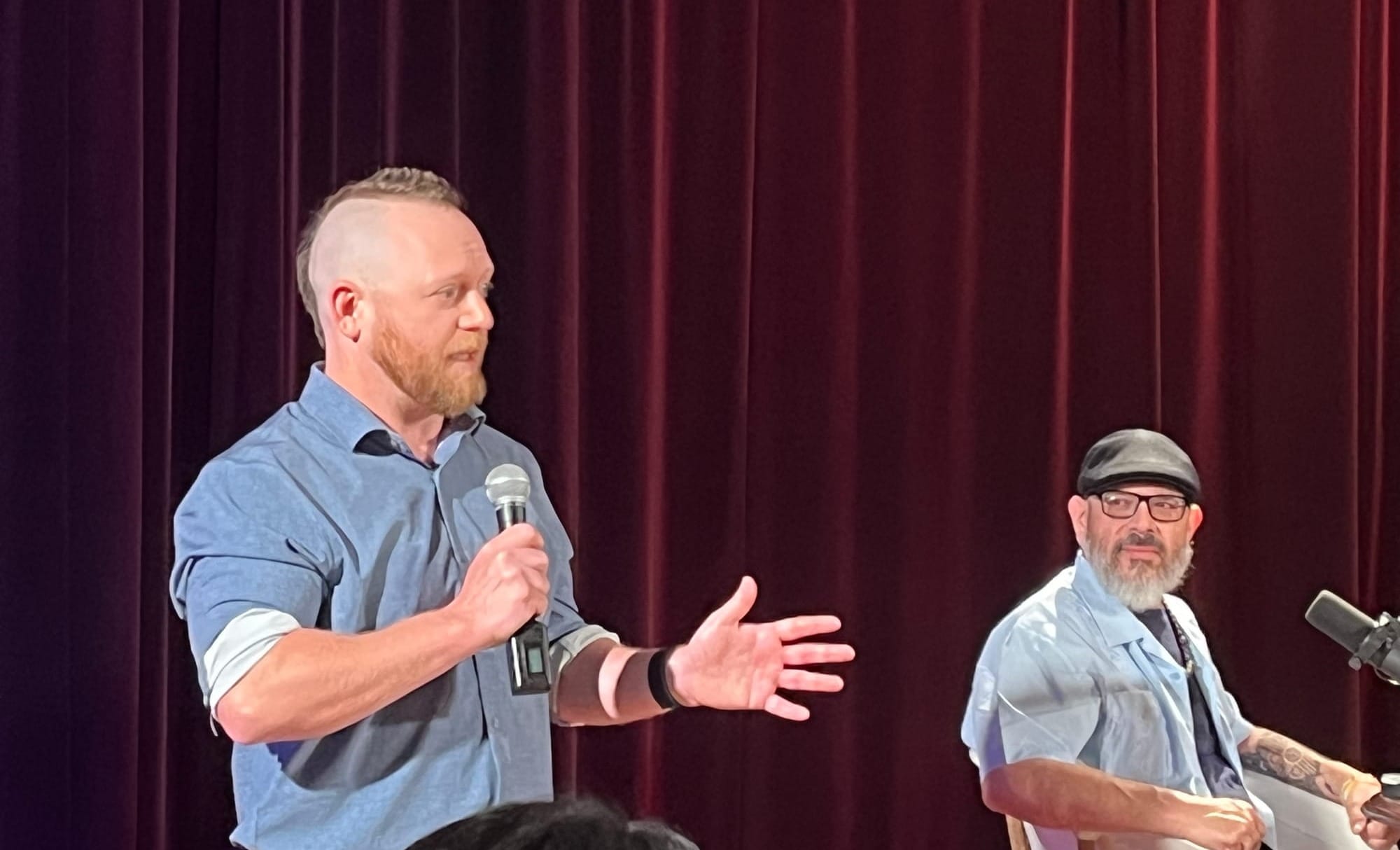 Micah McCoy speaks into a handheld microphone while introducing “Pass the Sopapillas” host Manuel González, who sits beside him onstage at KRWG Studios.