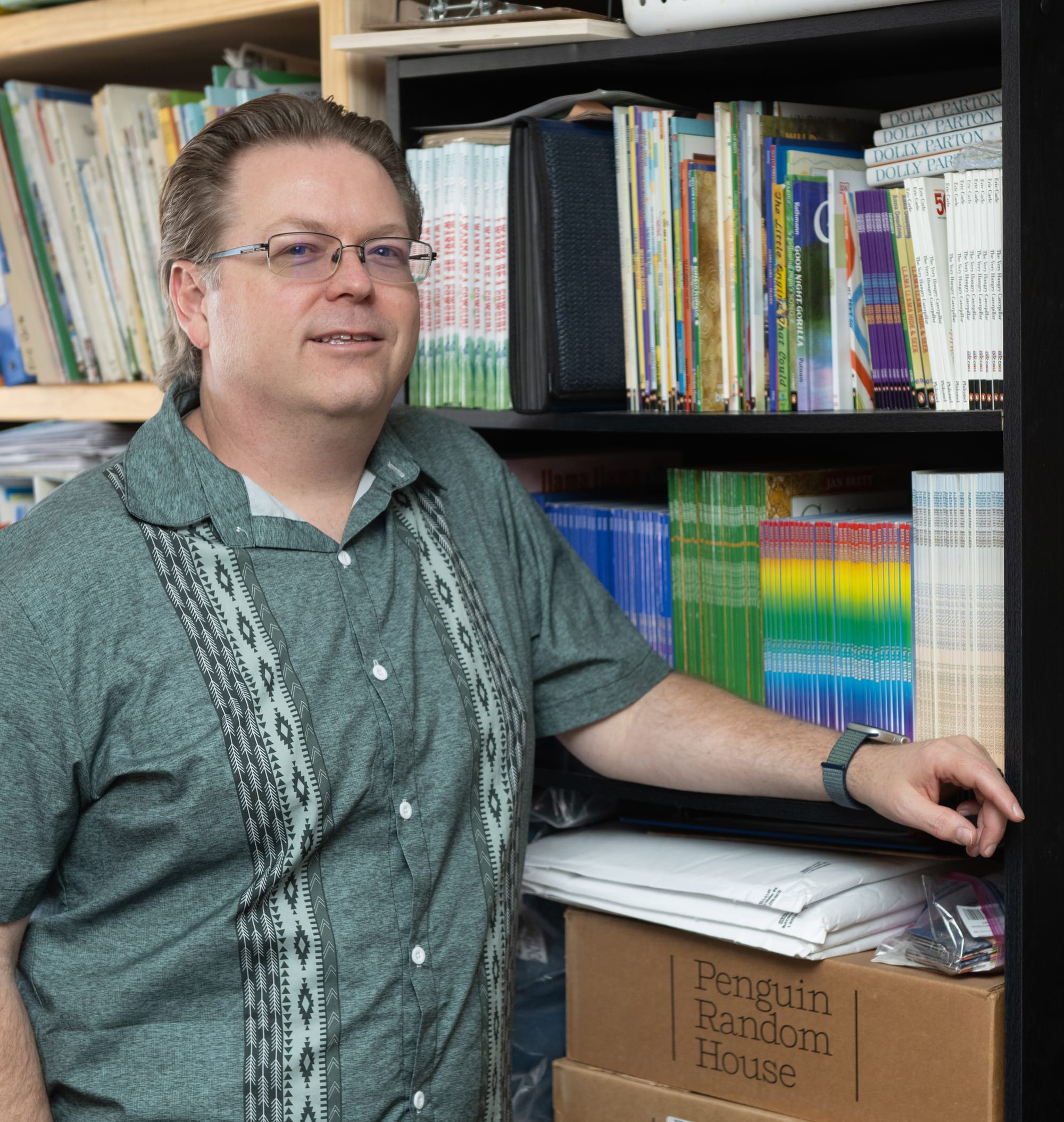 Ryal McMurry stands beside shelves of children’s books and boxes from Penguin Random House.