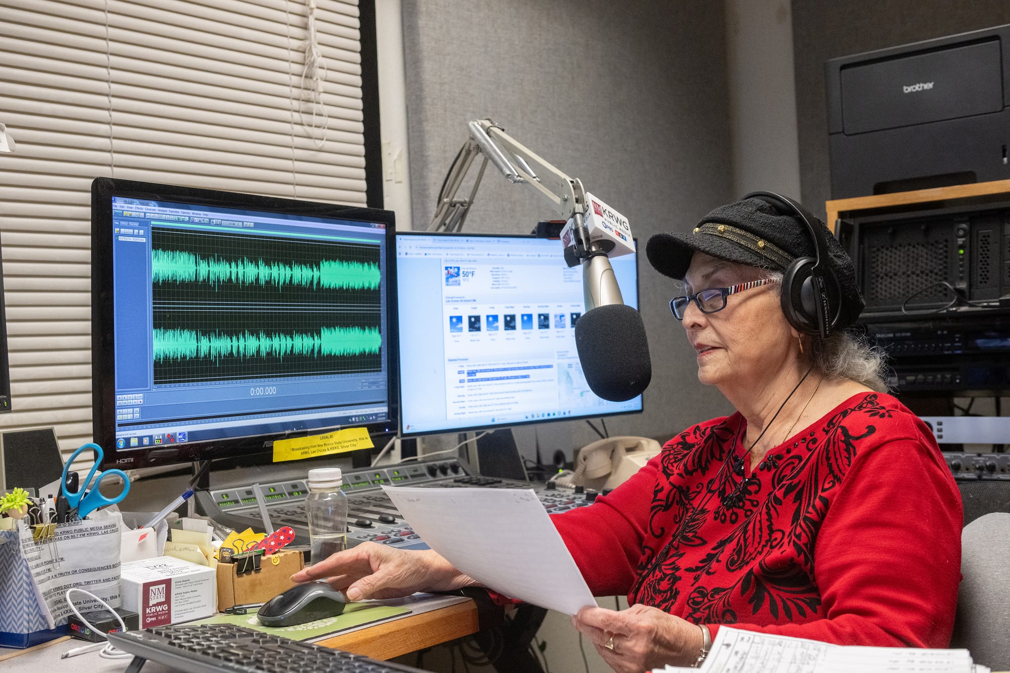 Emily Guerra works at a KRWG Public Media studio microphone while wearing headphones and reading from a sheet of paper.