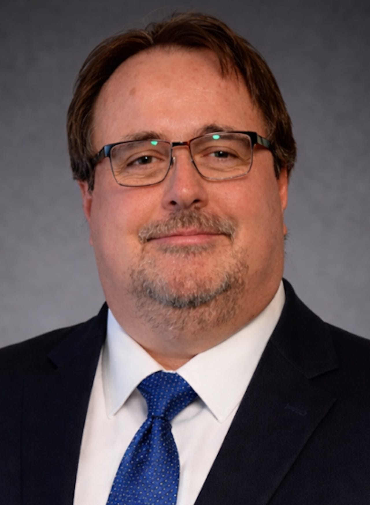 Moises M. Gutierrez wears glasses, a dark suit and a blue tie in a portrait against a gray background.