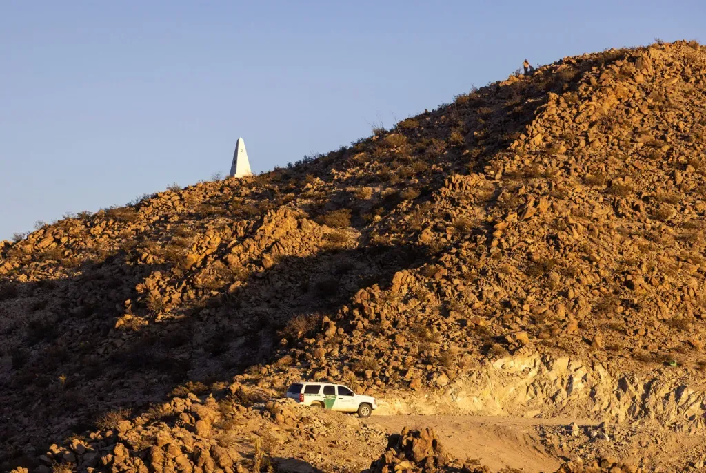 A Border Patrol vehicle sits on a rocky road below Mount Cristo Rey, where an obelisk marks the U.S.-Mexico border and a person stands on the ridgeline.