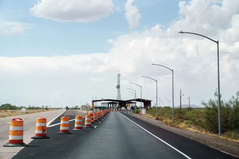 Orange-and-white traffic barrels separate lanes leading to a U.S. Customs and Border Protection checkpoint on Interstate 25 in southern New Mexico, beneath a partly cloudy sky.